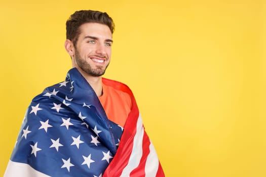 Proud caucasian man wrapping with a north america national flag in studio with yellow background