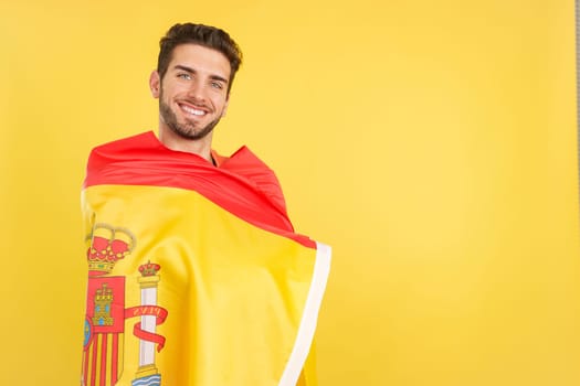 Happy caucasian man wrapping with a spanish national flag in studio with yellow background