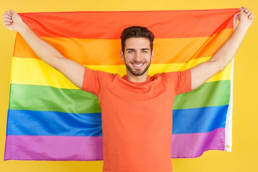 Happy caucasian man raising a lgbt rainbow flag in studio with yellow background