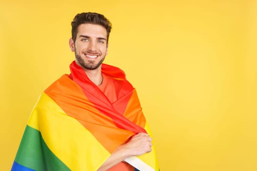 Happy caucasian man wrapped with a lgbt flag in studio with yellow background
