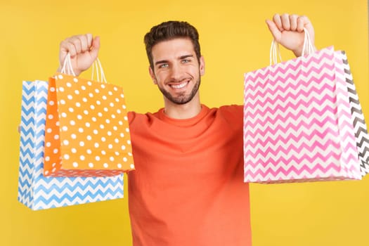 Caucasian man raising many shopping bags while smiling in studio with yellow background
