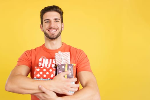 Happy caucasian man holing many colorful gifts in studio with yellow background