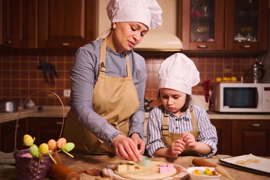 Delightful woman, loving mother and adorable baby girl, lovely daughter in white chef's hat and beige kitchen apron, putting cookie cutter on rolled dough, cooking together pastries for Easter holiday