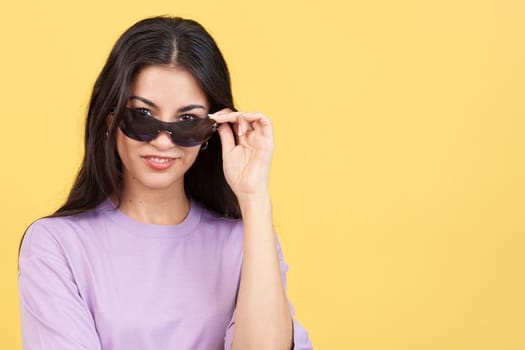 Hispanic woman lowering her sunglasses to see and look at the camera in studio with yellow background