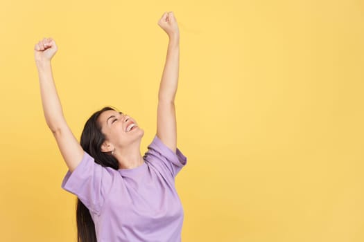 Happy caucasian woman celebrating while raising arms in studio with yellow background