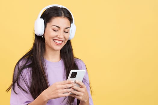 Caucasian woman listening to music with headphones and a mobile in studio with yellow background