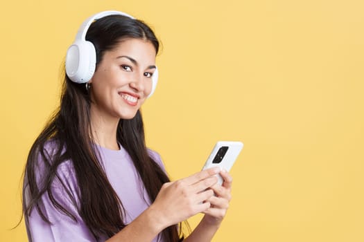 Caucasian woman looking at camera while listening to music with headphones and mobile in studio with yellow background
