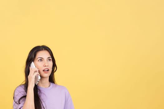 Surprised hispanic woman talking to the mobile in studio with yellow background