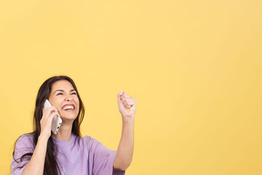 Happy woman celebrating while taking to the mobile in studio with yellow background