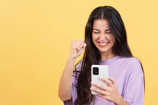 Excited woman celebrating while using the mobile phone in studio with yellow background
