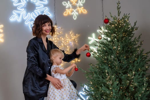 A mother with a 2-year-old daughter decorates the Christmas tree. Mom in a black suit, a girl in a white dress, her daughter hangs a red ball on the Christmas tree. Merry Christmas and New Year concept.