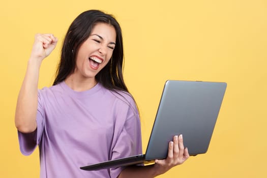 Caucasian woman celebrating while looking at the laptop in studio with yellow background