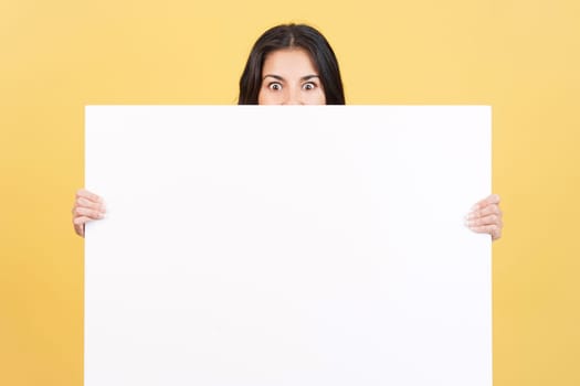 Surprised woman hiding behind a blank panel in studio with yellow background