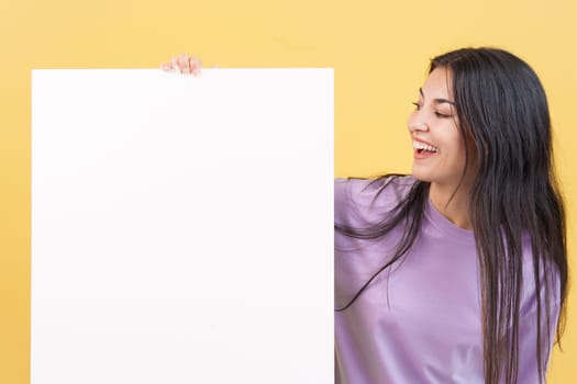 Happy caucasian woman looking and holding a blank panel in studio with yellow background