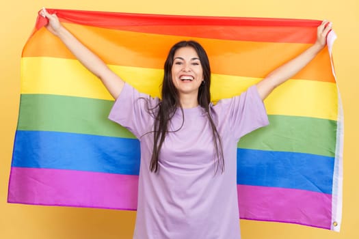 Happy caucasian woman raising a lgbt rainbow flag in studio with yellow background