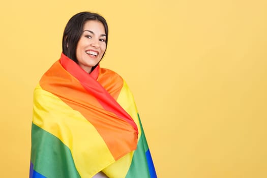 Happy caucasian woman wrapped with a lgbt flag in studio with yellow background
