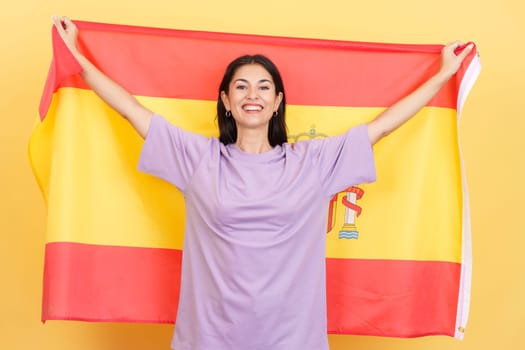 Happy caucasian woman smiling and raising a spanish national flag in studio with yellow background