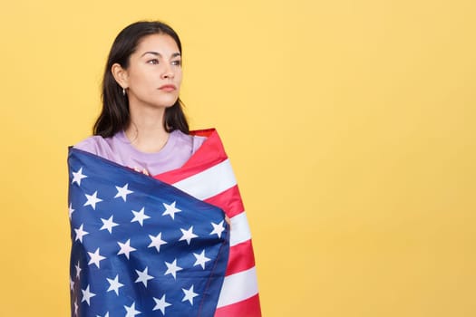 Proud and serious hispanic woman wrapping with a north america national flag in studio with yellow background