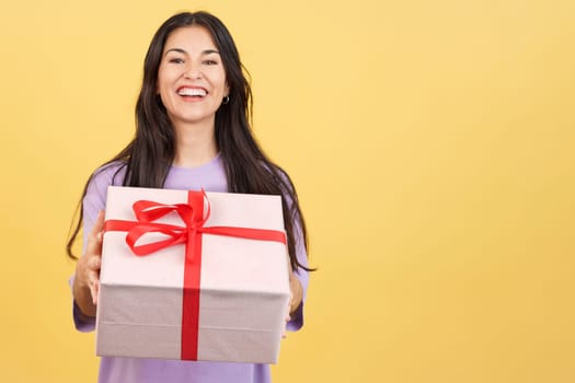 Happy woman looking at the camera giving a present in studio with yellow background