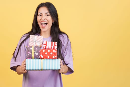 Happy caucasian woman holing many colorful gifts in studio with yellow background