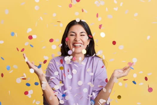 Happy woman surrounded by confetti flying in the air in studio with yellow background