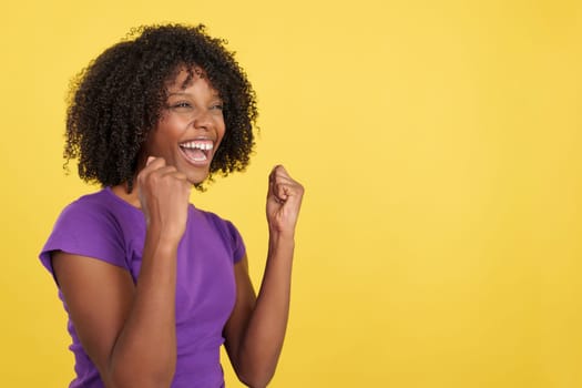 Happy woman with afro hair celebrating while raising fists in studio with yellow background