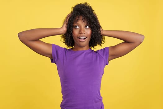 African-haired woman with hands on head expressing surprise in studio with yellow background