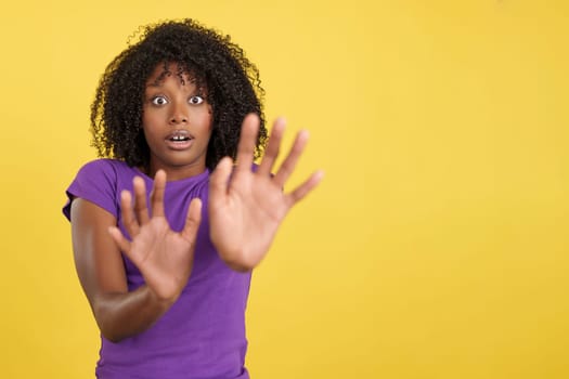 Woman with African hair expressing fear while shielding herself with her hands in studio with yellow background