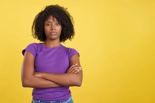 Angry woman with afro hair standing with arms crossed in studio with yellow background
