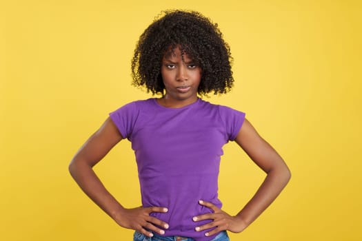 Woman with afro hair looking furious at camera with hands on hips in studio with yellow background