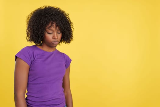 Woman with afro hair looking down with sad expression in studio with yellow background