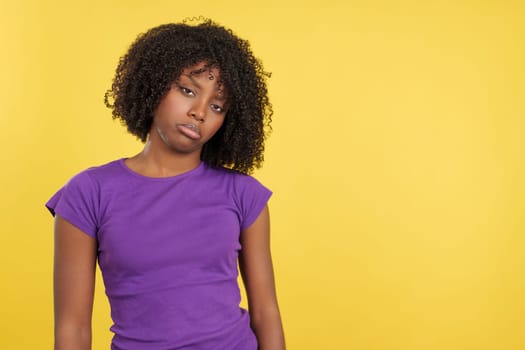 Sad woman with afro hair standing looking at camera in studio with yellow background