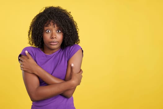 Woman with African hair hugging herself gesturing coldness in studio with yellow background