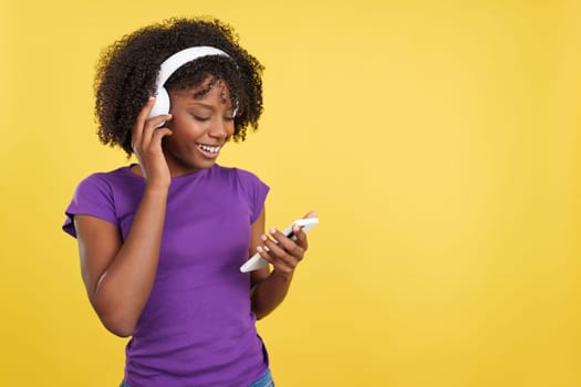 Afro woman listening to music with mobile phone and headphones in studio with yellow background