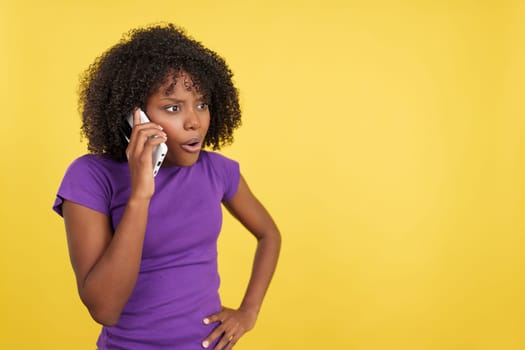 Woman with afro hair talking to the mobile with angry expression in studio with yellow background