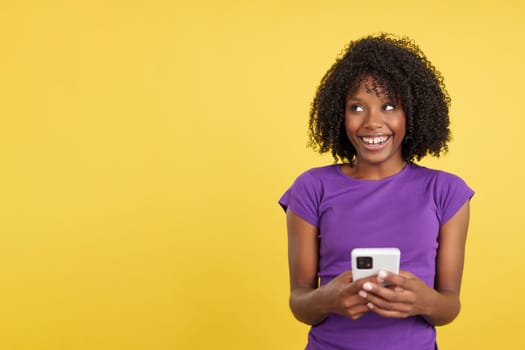 Happy woman with afro hair looking up while using a mobile in studio with yellow background