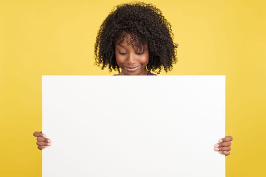 Woman with afro hair looking down to a blank board that she is holding in studio with yellow background