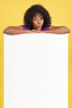 Surprised afro woman above a blank panel in studio with yellow background