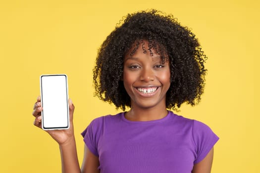 Woman with afro hair smiling at the camera holding a mobile with a white screen in studio with yellow background