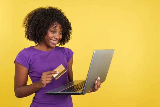 Woman with afro hair shopping online with a laptop and a credit card in studio with yellow background