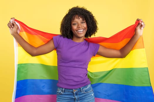 Happy woman with afro hair smiling while looking at camera and raising a lgbt rainbow flag in studio with yellow background