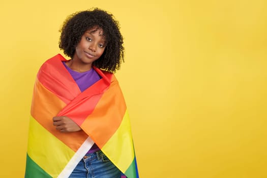Woman with afro hair wrapped with a lgbt rainbow flag in studio with yellow background