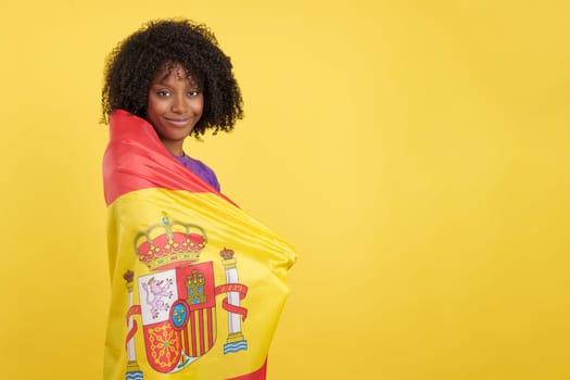 Migrant woman with afro hair wrapped with a Spanish flag in studio with yellow background