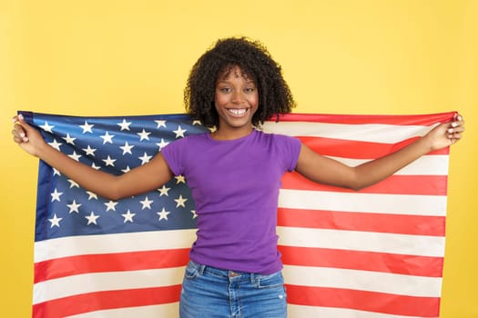 Happy woman with afro hair raising a United States flag in studio with yellow background