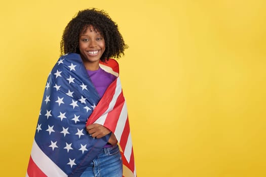 Happy woman with afro hair wrapping with a united states flag in studio with yellow background