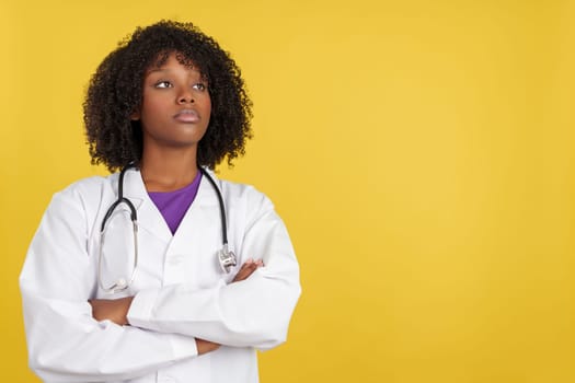 Proud afro female doctor looking up with arms crossed in studio with yellow background