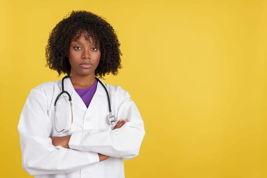 Serious afro doctor with stethoscope looking at camera with arms crossed in studio with yellow background