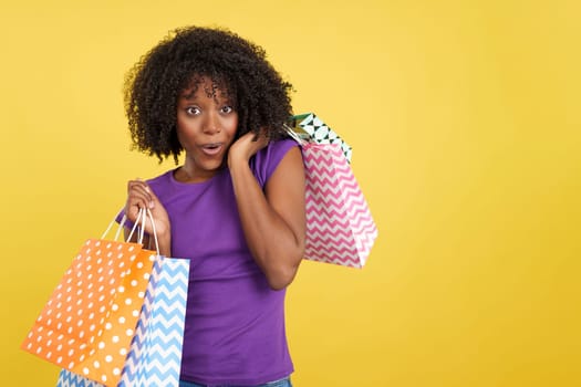 Greedy woman with afro hair holding many shopping bags in studio with yellow background