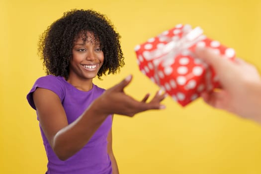 Cool woman with afro hairstyle smiling while receiving a gift in studio with yellow background