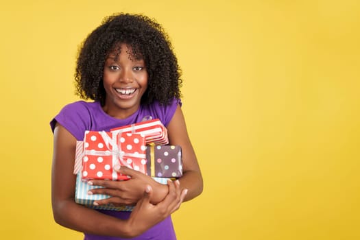 Happy woman with afro hairstyle smiling at the camera holding gifts in studio with yellow background